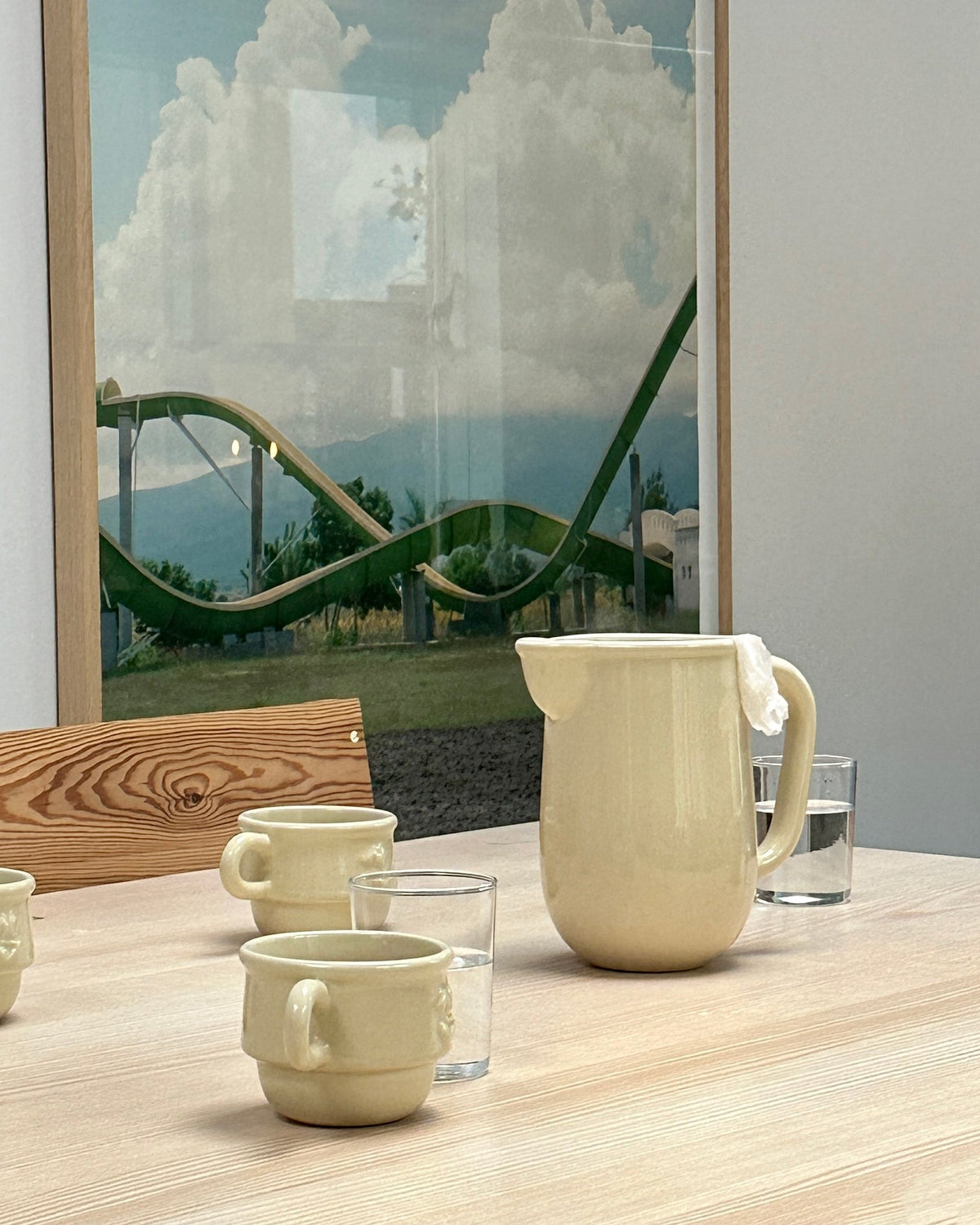 Beige Kantine stoneware pitcher by NIKO JUNE arranged with matching ceramic cups and Soft Serve drinking glasses on a wooden table.