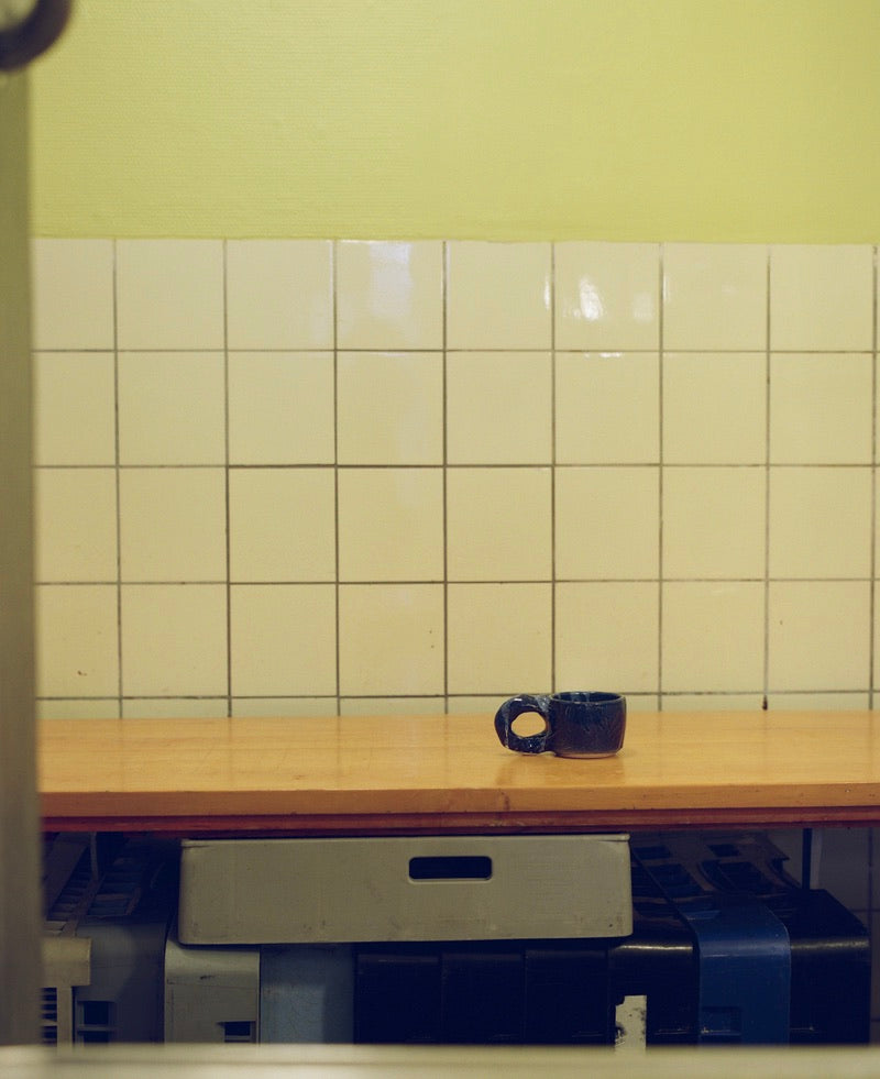 Dark Blue Studio Cup by NIKO JUNE displayed against a yellow tiled wall in a minimalist interior.