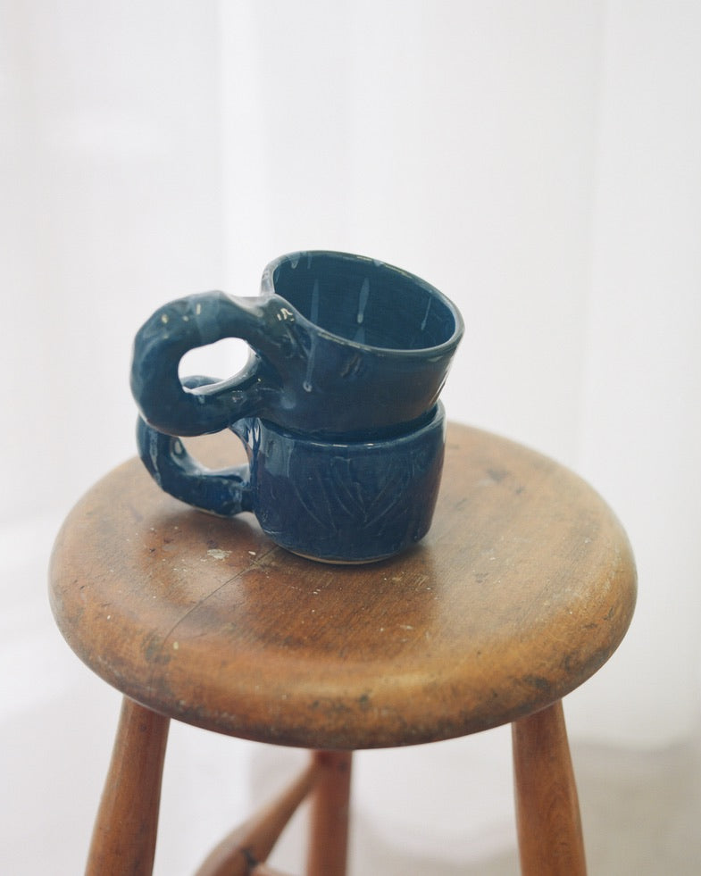 Two handmade Studio Cups in Dark Blue by NIKO JUNE stacked on a wooden stool, showing their sculptural handles and glazed stoneware surfaces.