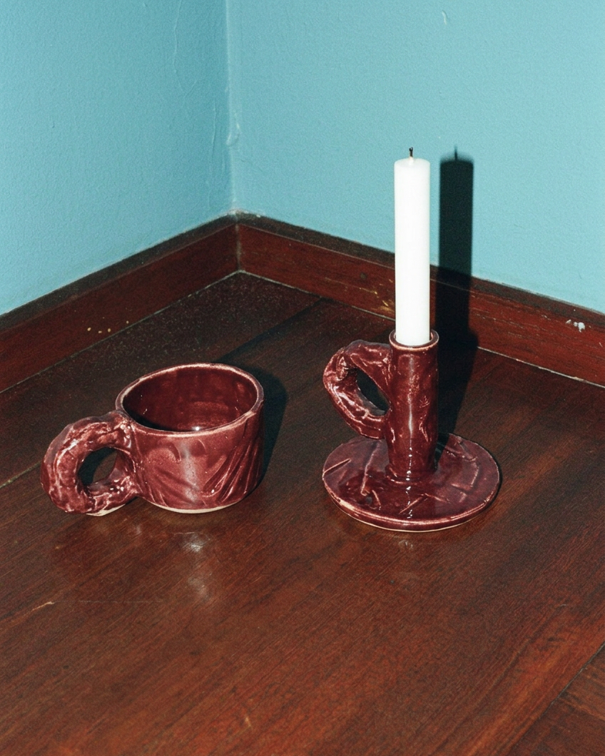 Studio Candlestick Burgundy by NIKO JUNE holding a dark candle on a wooden table, alongside a matching burgundy Studio Cup against a turquoise wall.