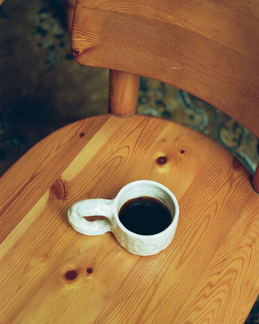 Studio Cup White by NIKO JUNE photographed from above on a wooden chair, showing its sculptural handle and white ceramic glaze.